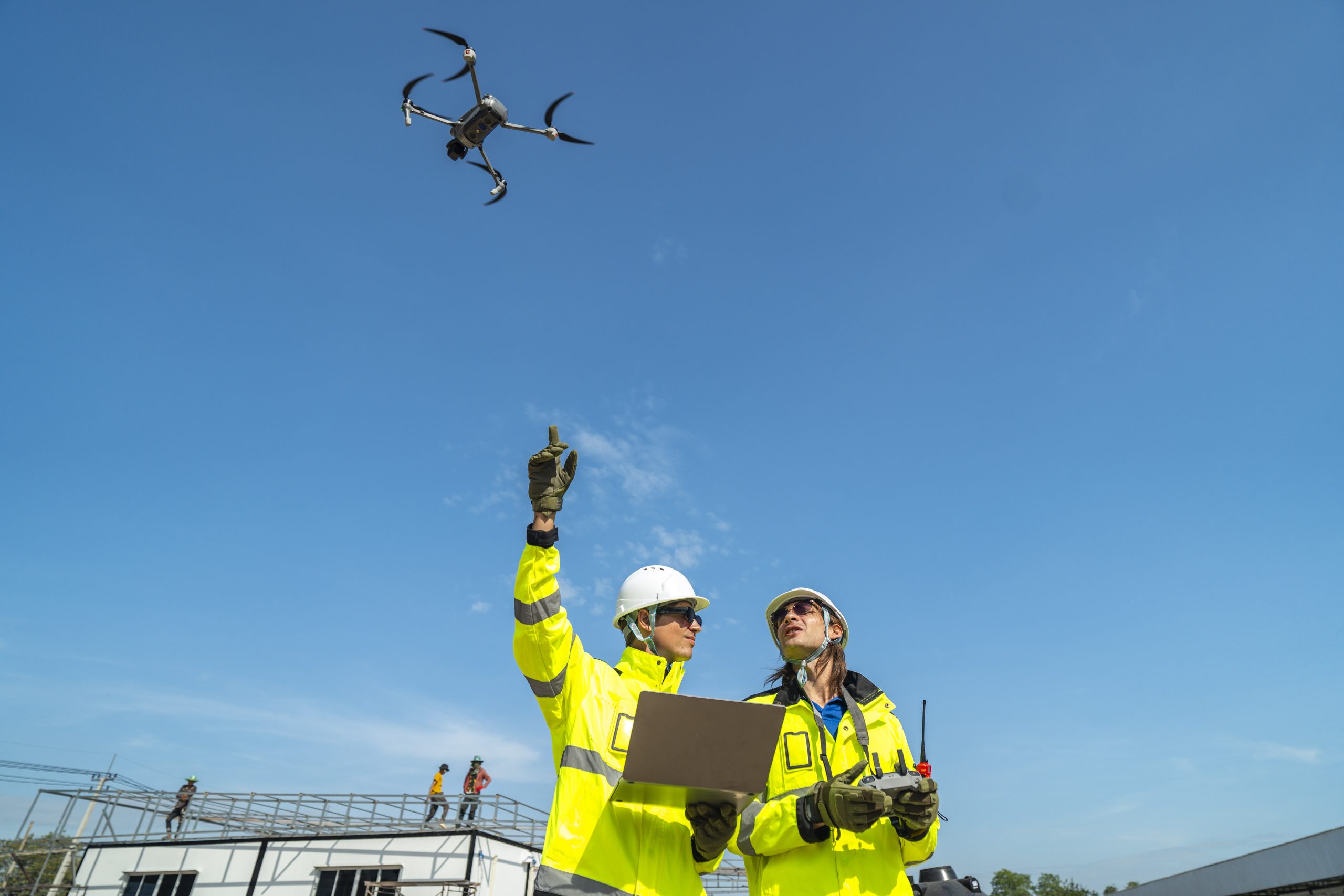 Two engineers in high visibility jackets and helmets observing a drone flying over a construction site, with machinery and modular buildings, highlighting technology and site monitoring.
